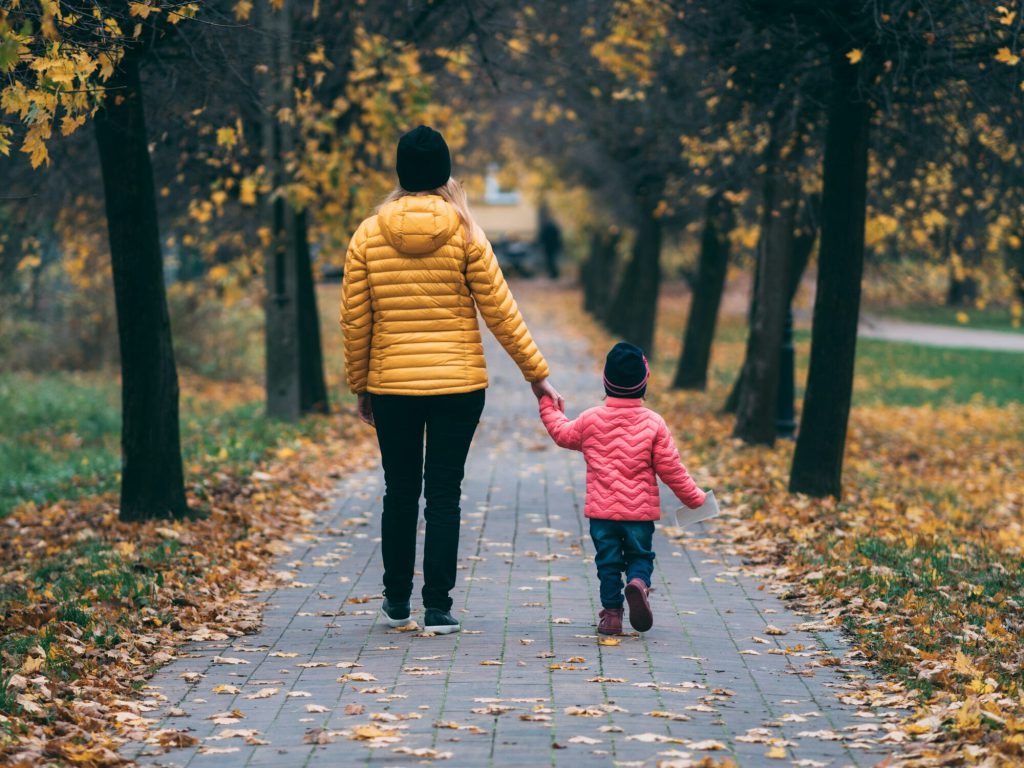 A parent and young child walking hand in hand along a leaf-covered park path